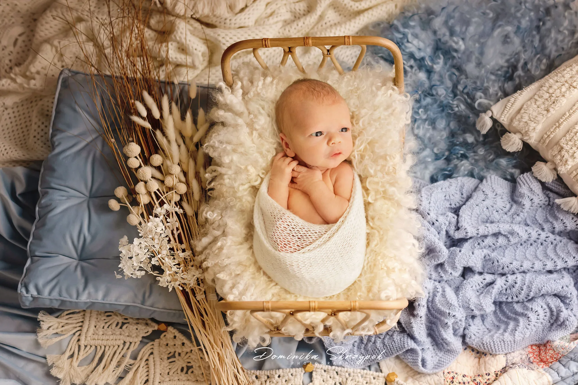 Newborn wrapped in white knit, sleeping in a wicker basket among wraps and fabrics in a Warsaw studio