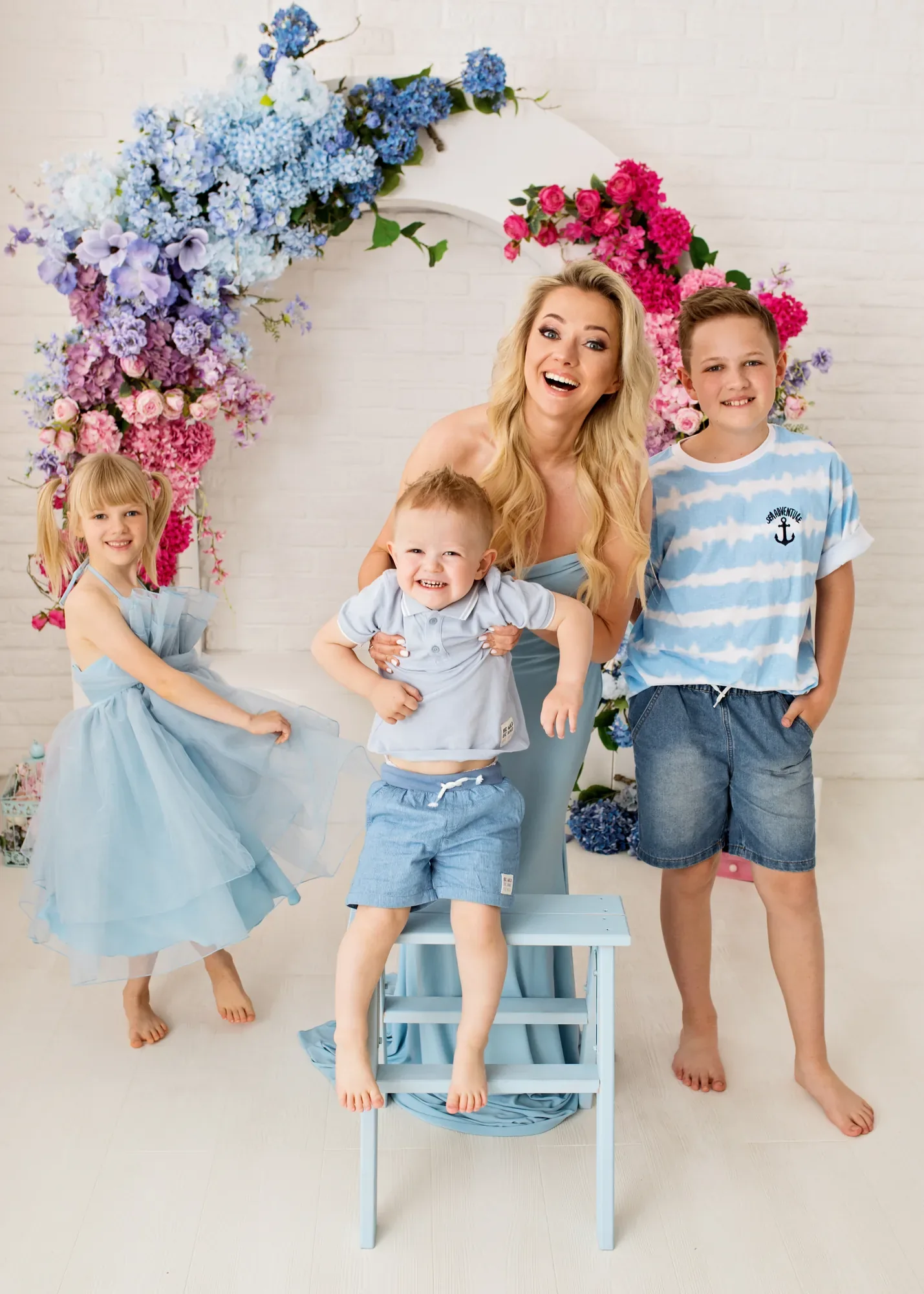 Joyful family session in pale blue outfits — mum with her three children by a floral arch in a studio in Warsaw