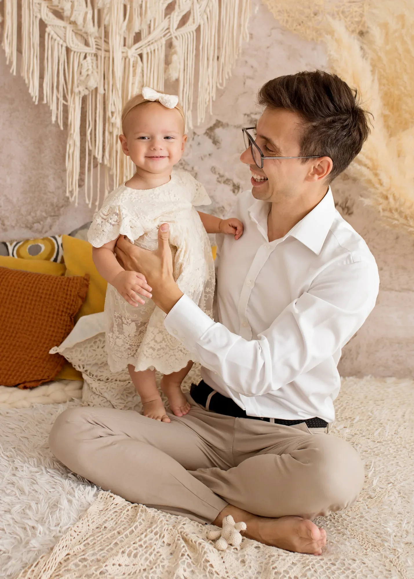 Boho family session — smiling dad with his daughter in a lace dress on a light rug in a studio in Warsaw